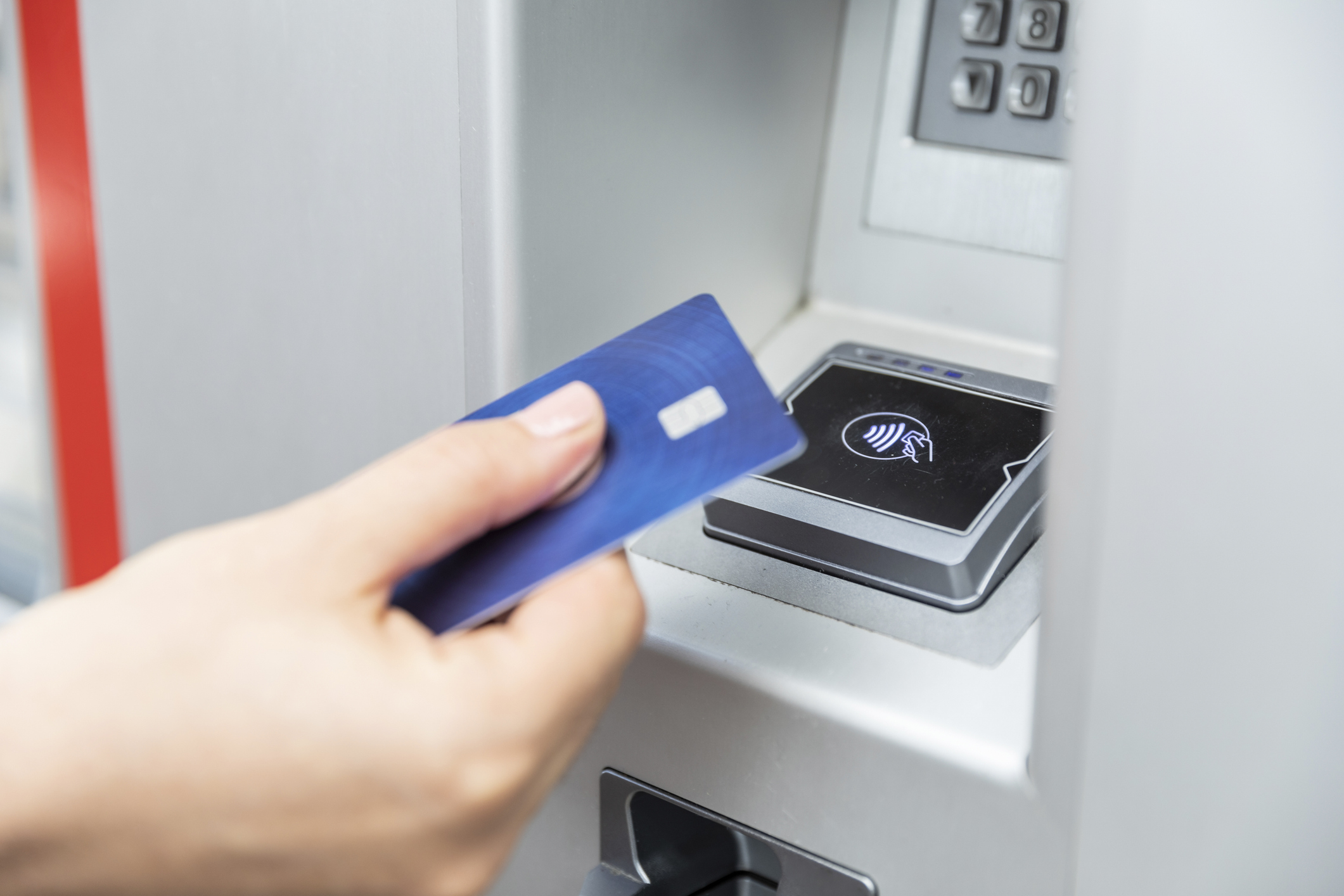 A person holding a blue contactless credit card near an ATM reader to make a tap payment, illustrating modern banking technology and secure contactless transactions.